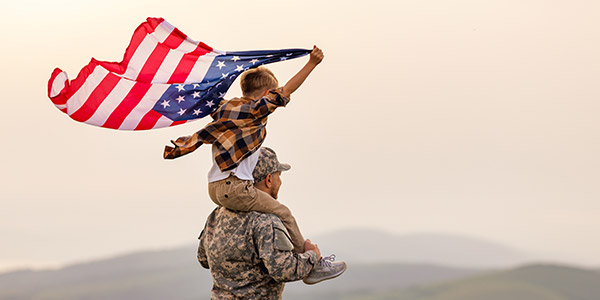 A white man in an American military uniform stands with a young boy, holding an American flag, on his shoulders