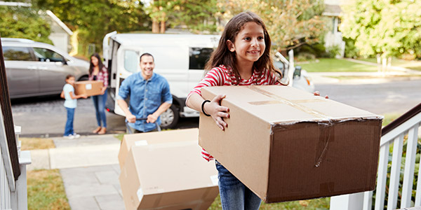 A little smiling Hispanic girl carries a moving box into a home while her parents walk behind her