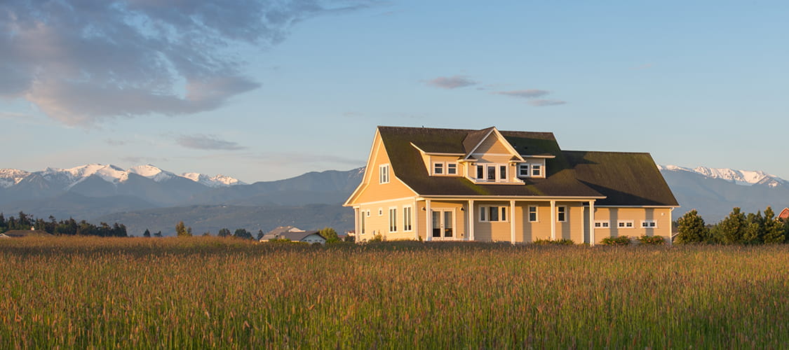 A large two story house sits on open grass land with mountains in the background