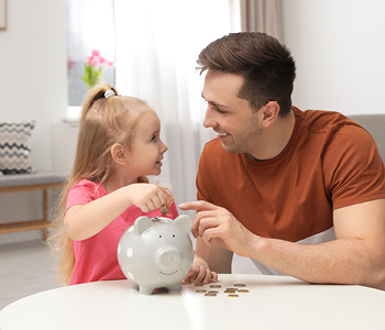 Punčka z očetom sedi za mizo in daje kovanec v keramični hranilnik - pujska. Slika simbolizira učenje varčevanja.