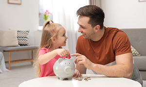 Punčka z očetom sedi za mizo in daje kovanec v keramični hranilnik - pujska. Slika simbolizira učenje varčevanja.