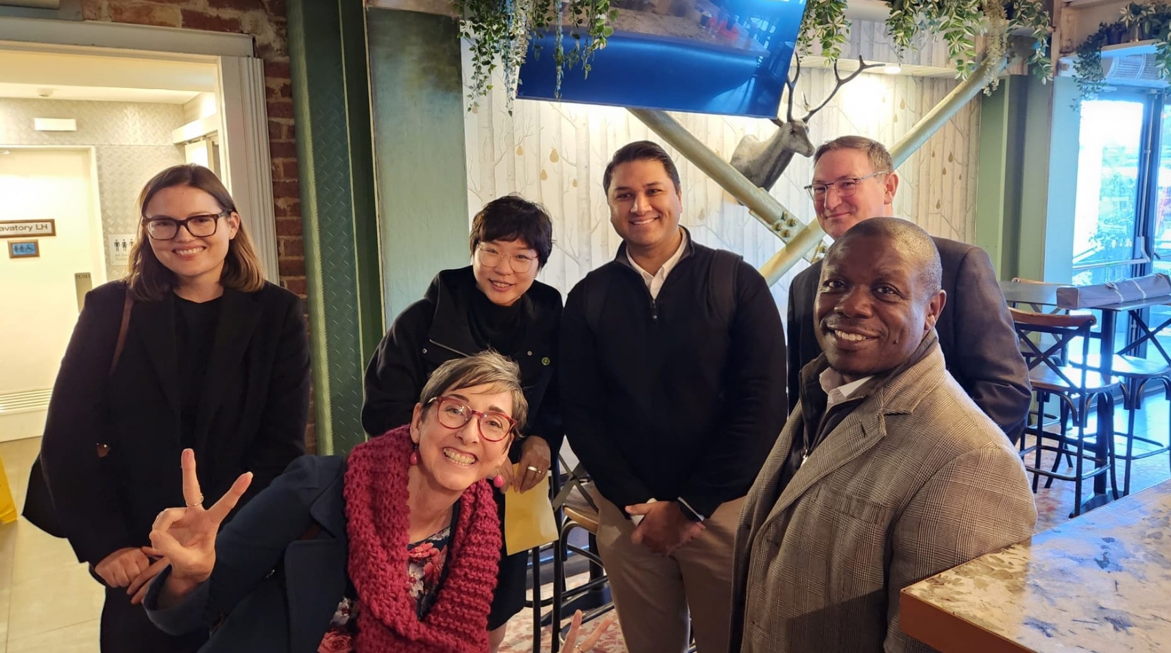 Six people are gathered for a photo in a casual, brightly lit cafe or restaurant setting. One woman in the foreground is kneeling and smiling, holding up a peace sign. The other five people, two women and three men, are standing behind her, all smiling for the camera. The background includes decorative elements like a mounted deer head and greenery.