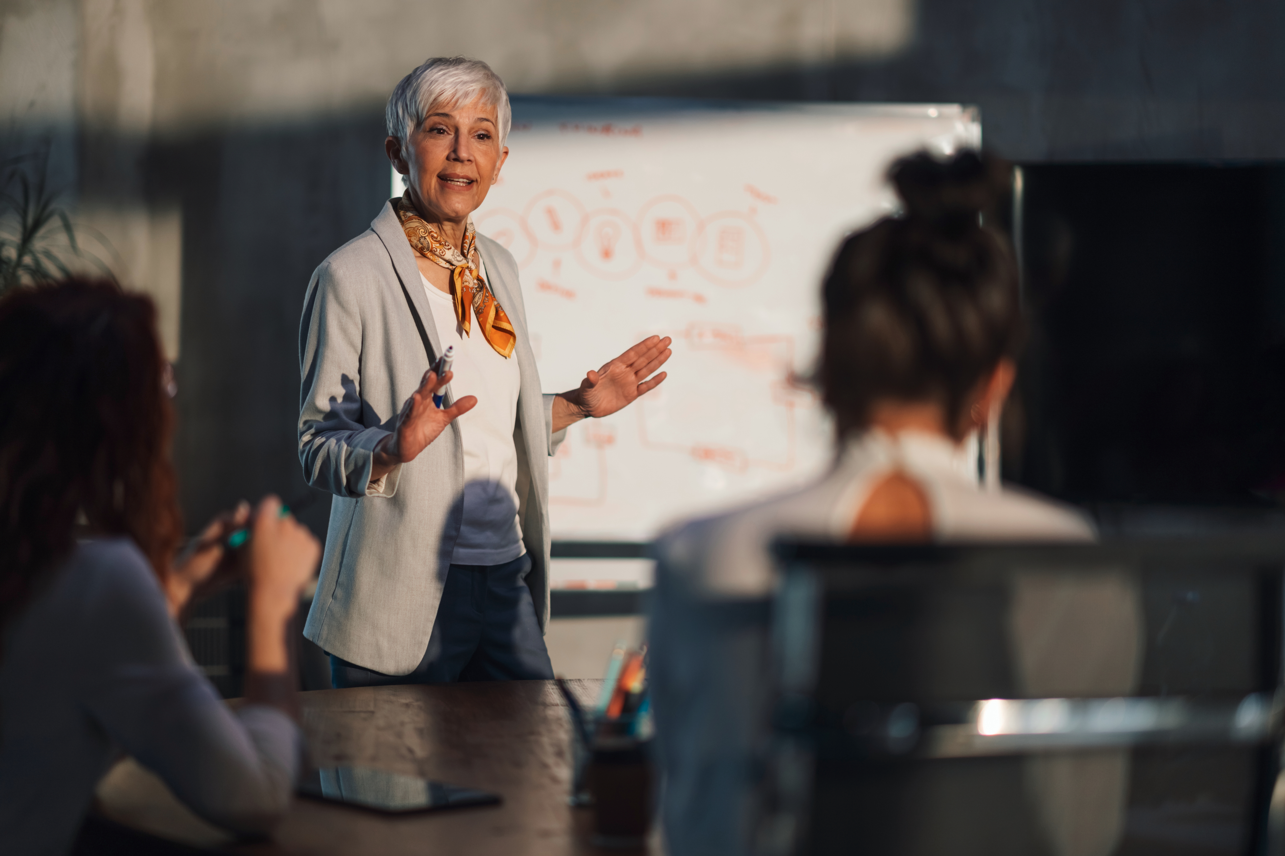 Confident senior businesswoman giving a presentation to colleagues in front of a whiteboard during a meeting.