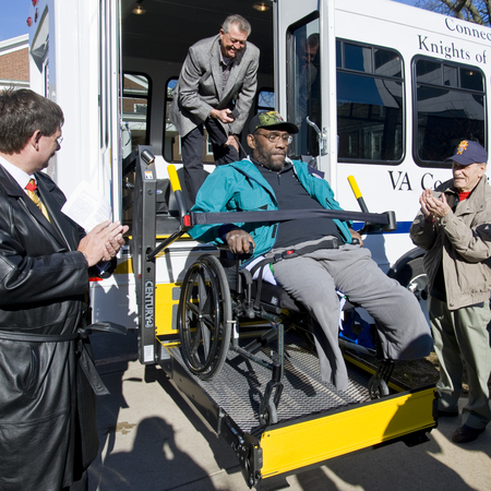 Knights assit a wounded veteran in a wheelchair off a VA transportation bus.