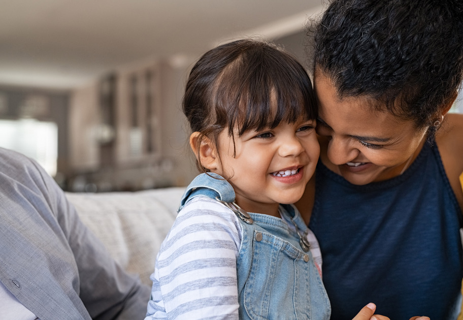 Young girl with brown hair and a front fringe smiles as her mum gives her a cuddle on the couch.