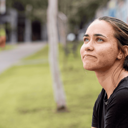 Young woman sitting outdoors on a bench, wearing a black shirt, looking upward with a thoughtful expression, with a mural wall and greenery in the background.
