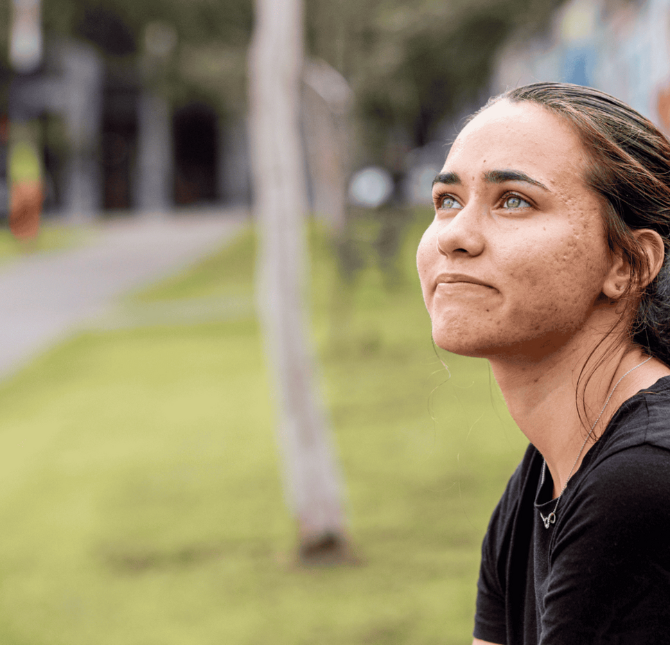 Young woman sitting outdoors on a bench, wearing a black shirt, looking upward with a thoughtful expression, with a mural wall and greenery in the background.