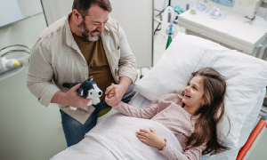 Srečen trenutek v bolnišnici, deklica leži v postelji, oče pa jo drži za roko, vesel okrevanja. Oba se smejita.