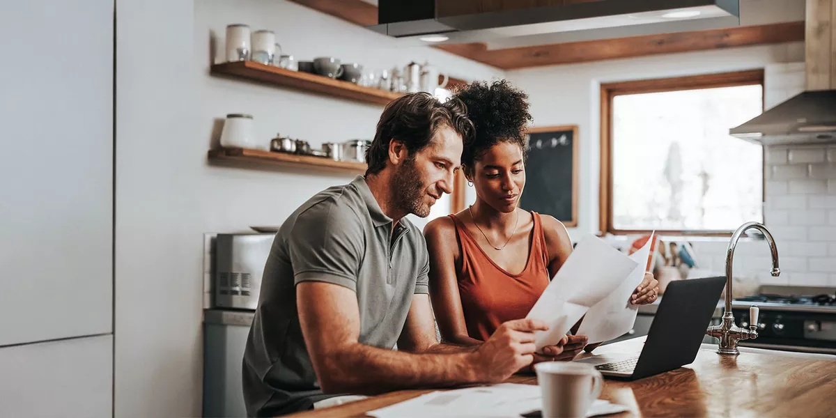 Two people seated at a kitchen counter reviewing papers beside an open laptop, with shelves, mugs, and a window visible in the background.