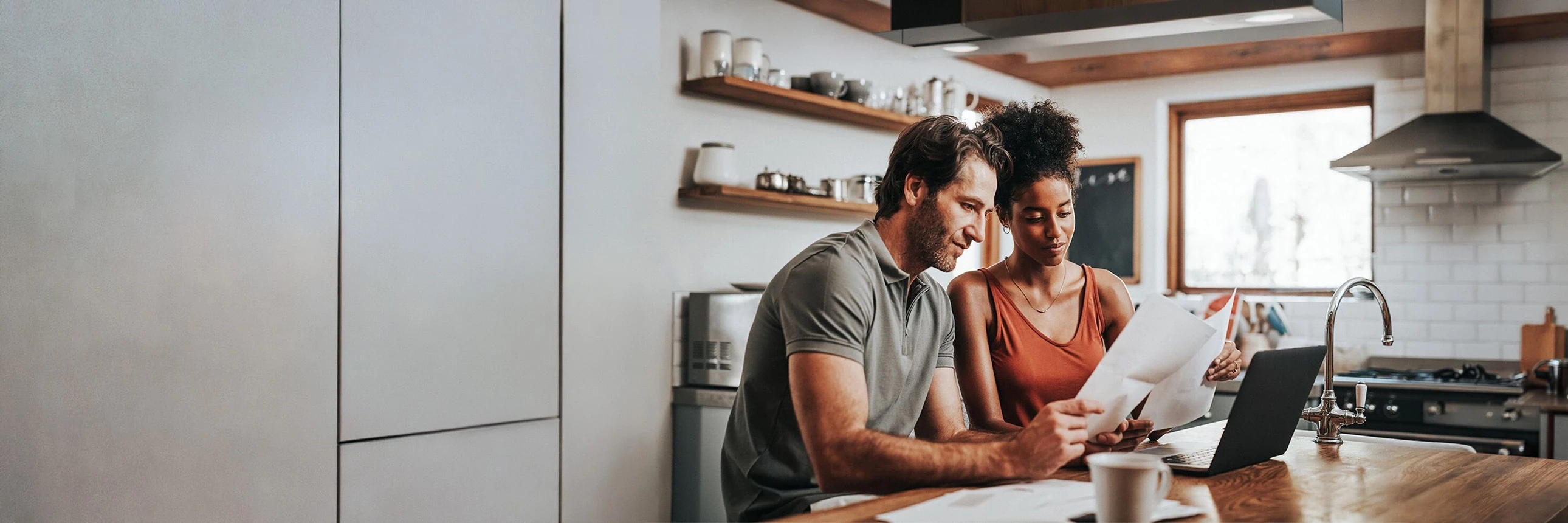 Two people seated at a kitchen counter reviewing papers beside an open laptop, with shelves, mugs, and a window visible in the background.