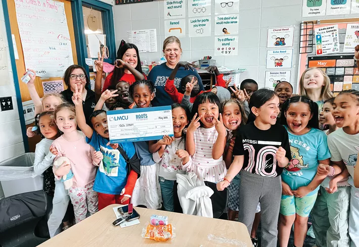 Group of children and two adults posing together in a classroom, with several children holding a large LMCU donation check.
