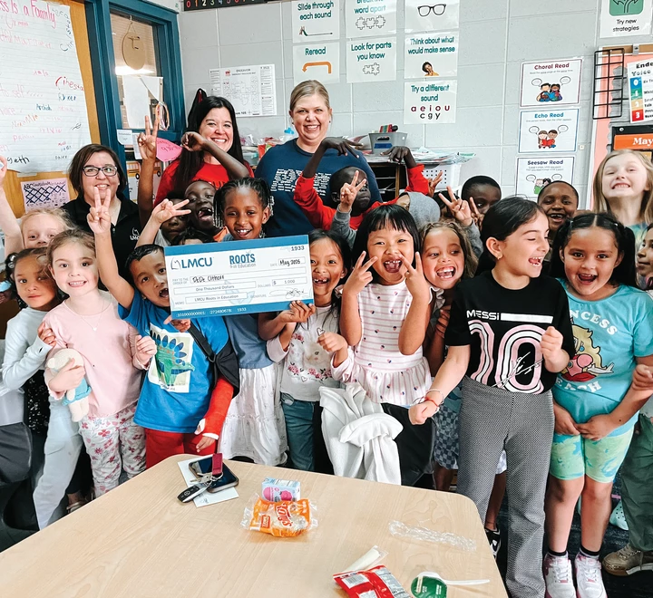 Group of children and two adults posing together in a classroom, with several children holding a large LMCU donation check.