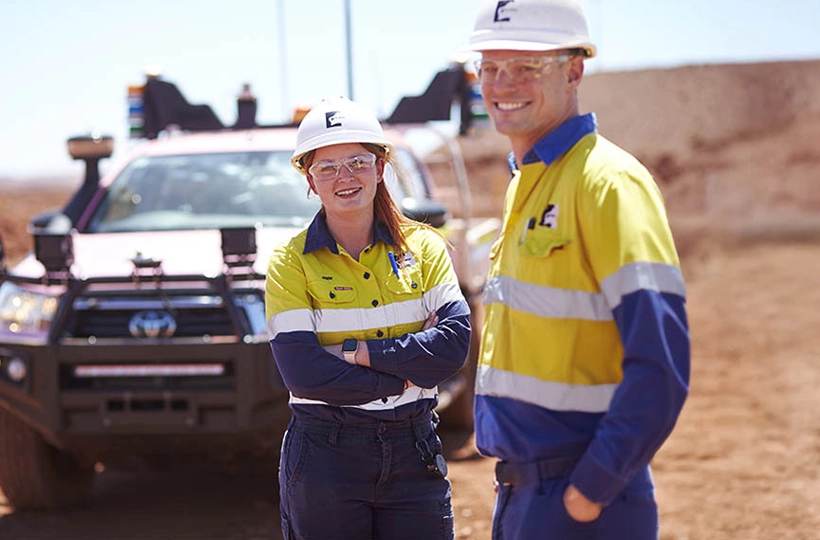 The image shows two workers in high-visibility clothing and helmets standing in front of a utility vehicle in a desert or mining area. The left side of the image features a map highlighting various locations and cable routes, including Darwin-Jakarta-Singapore Cable, Australia-Singapore Cable, Port Hedland, Project Horizon, Geraldton, and Perth. This image combines elements of fieldwork with geographical data representation.