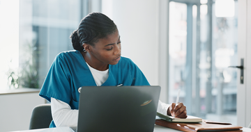 A female healthcare professional taking notes while working on a laptop in a clinical setting.