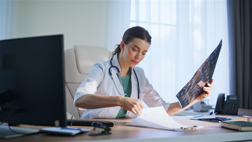 A female healthcare professional sitting at a desk reviewing medical scans and paperwork in a bright office. 