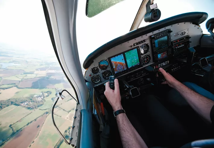 Pilot in cockpit flying over rural farmland. Photo is taken behind the pilot’s shoulder looking out the window. 