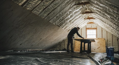 Wide shot of an attic renovation, man in background. Insulation is visible; plywood is on the bottom of the walls. 