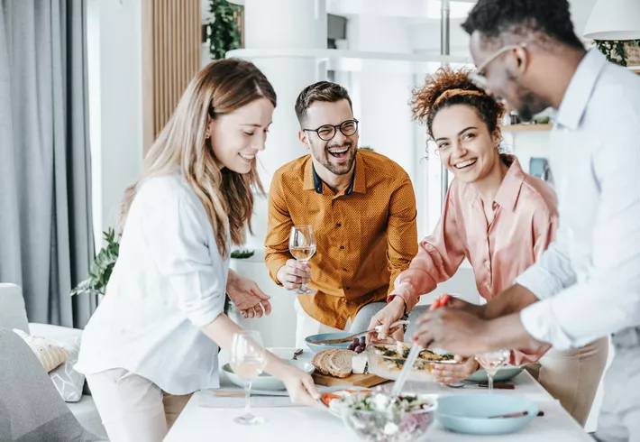 Four adults gathered around a dining table in a bright home kitchen, serving themselves food and holding drinks, with dishes like salad and bread spread across the table. 