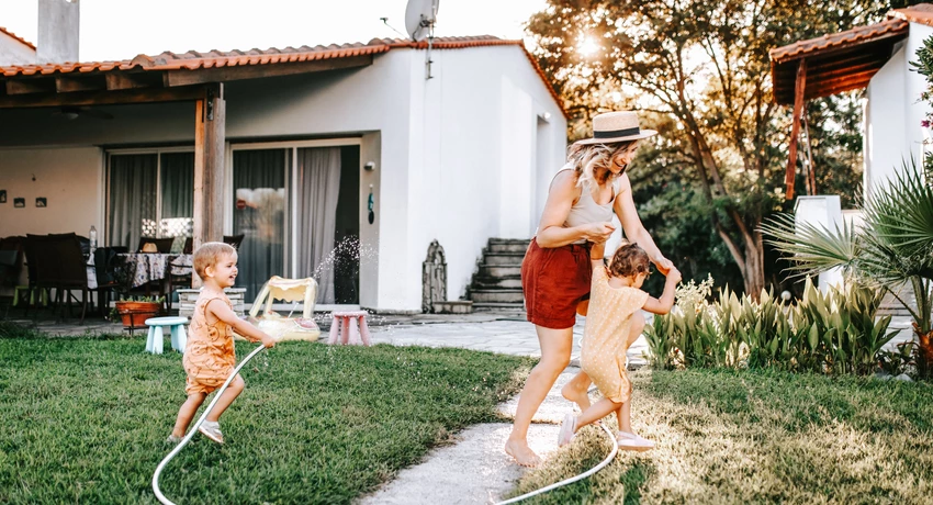 Mother playing in yard being sprayed with hose by her two young children 
