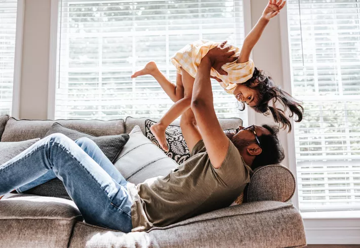 Person relaxing on a couch while lifting a child overhead in a bright living room with large windows.