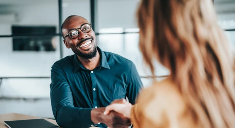 Two people shaking hands across a desk during an office meeting.